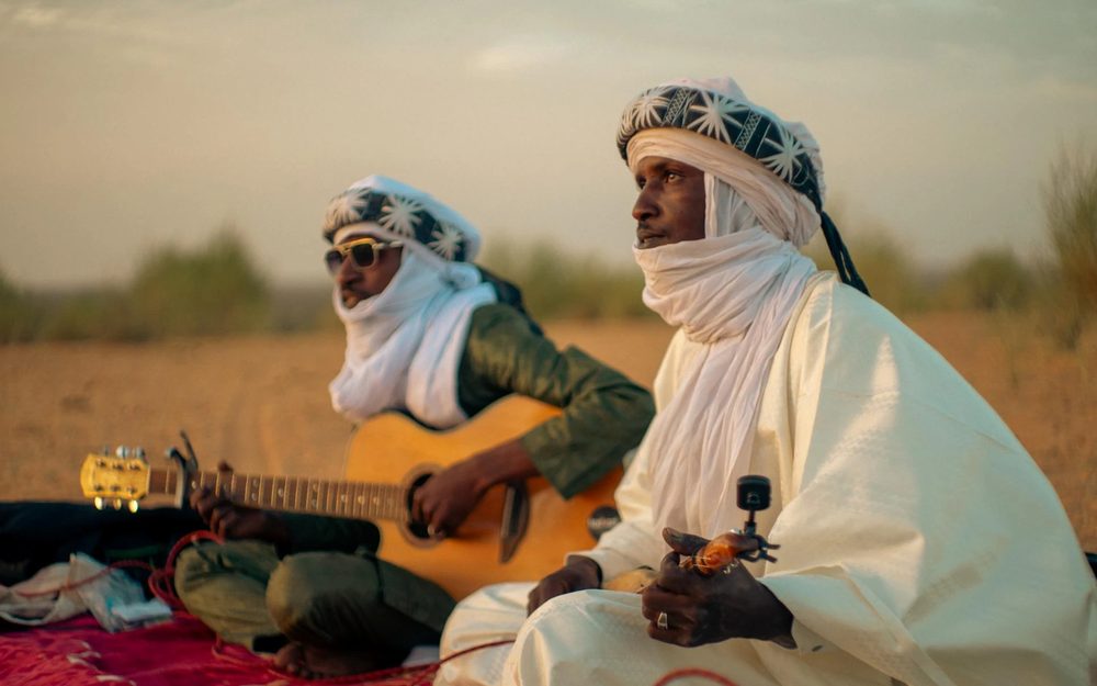 Two musicians in white robes and turbans sit in the desert, one playing guitar