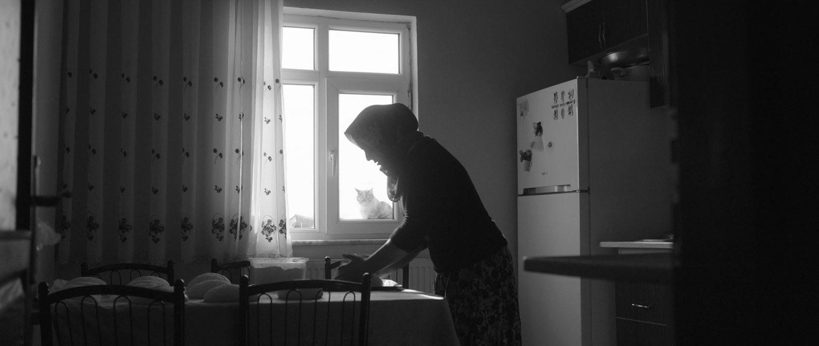 A woman shapes dough at a kitchen table while a cat looks in through the window