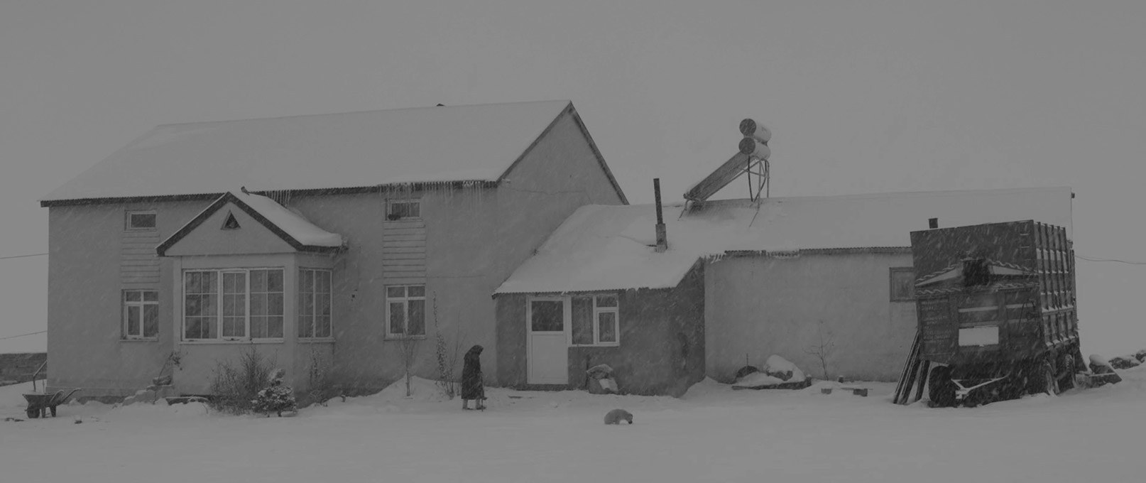 A farmhouse in heavy snow with an old woman walking past