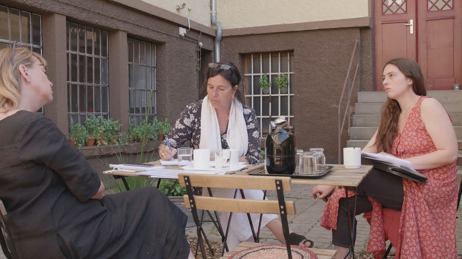 Three women sit at a table in a courtyard with papers and coffee cups