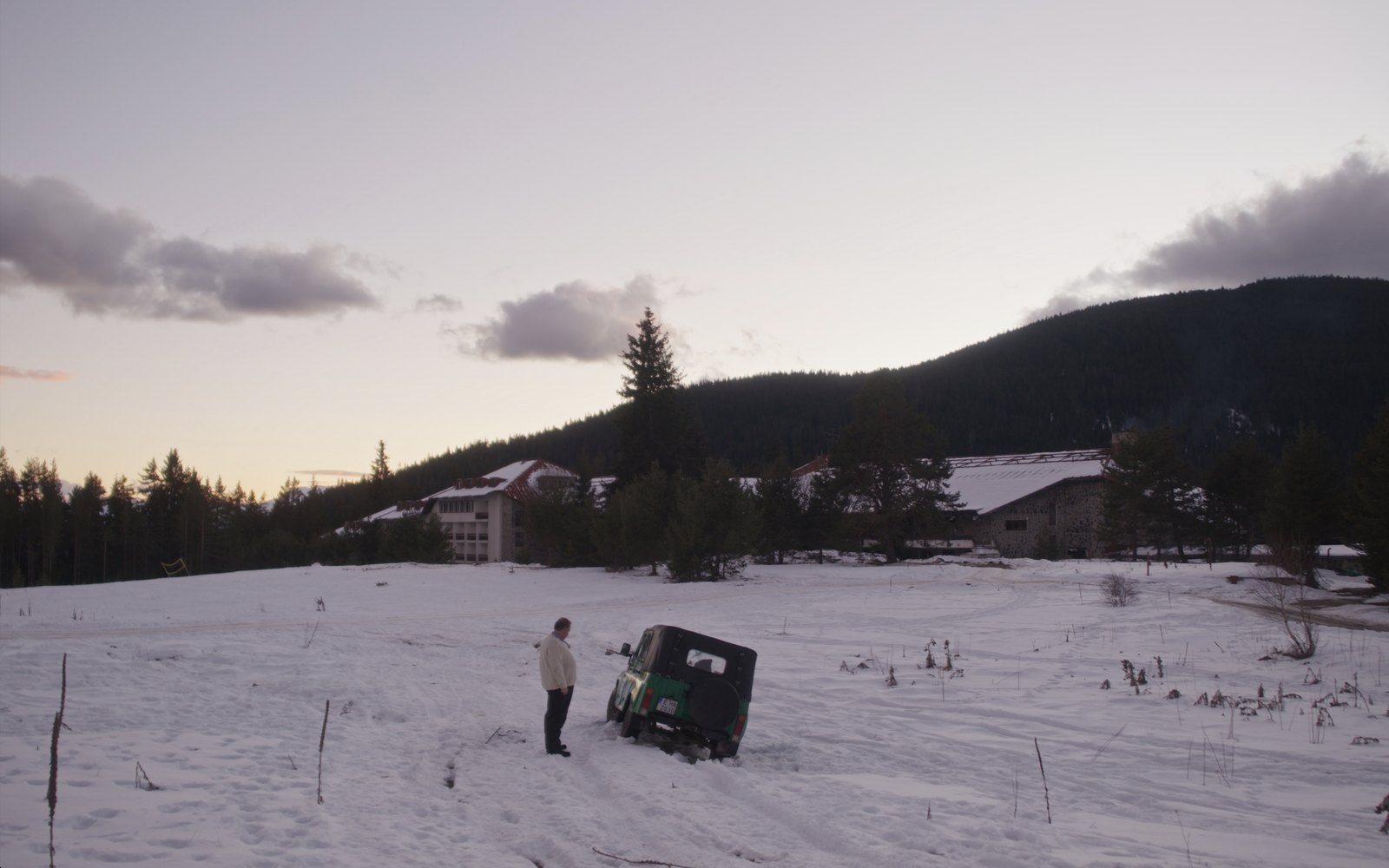 A man stands beside a jeep stuck in the snow in front of an abandoned mountain hotel
